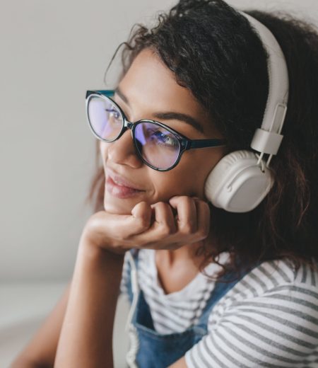 Close-up photo of dreamy girl with light-brown skin thinking about something pleasant while enjoying music. Indoor portrait of brunette young woman sitting with cup of coffee propping up head.
